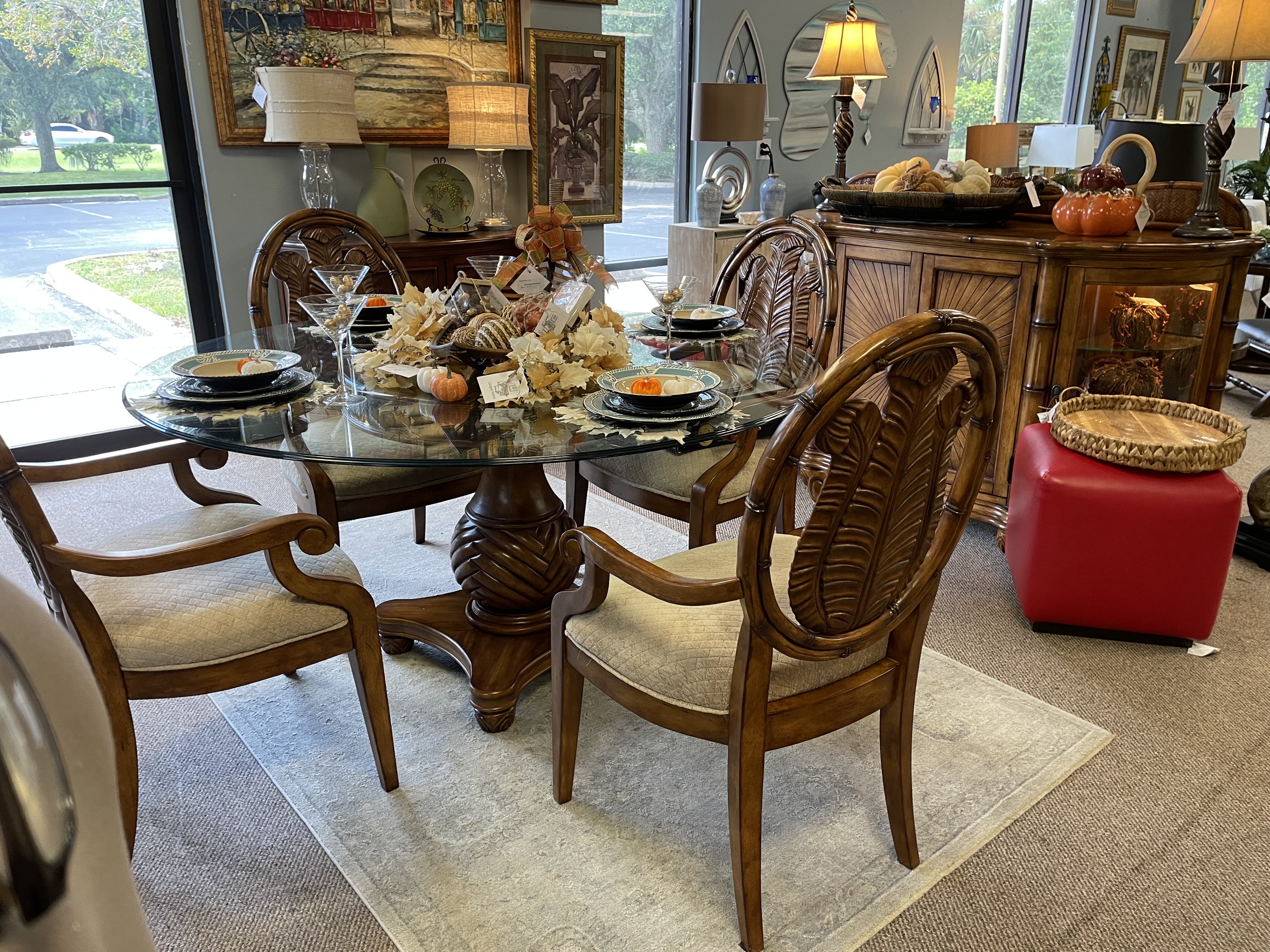 Elegant round glass 4-person dining table with ornate wooden chairs and beige seat cushions with place settings and centerpiece.